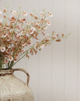 The Faire Wild Orchid Spray features delicate pink and white faux flowers with slender green stems, arranged in a rustic ceramic vase and set against a light, vertically paneled background for an airy home decor accent.