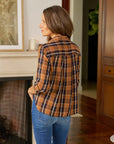 A woman with long brown hair wearing a Frank & Eileen Silvio Untuckable Shirt and blue jeans stands indoors, facing away and looking to the side, near a fireplace and wooden furniture.