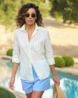 A person with curly hair and sunglasses walks confidently by a pool, wearing the relaxed fit Eileen S25 shirt by Frank & Eileen, made from Italian cotton, paired with blue shorts. They hold a straw hat with greenery and a sunlit pathway in the background.