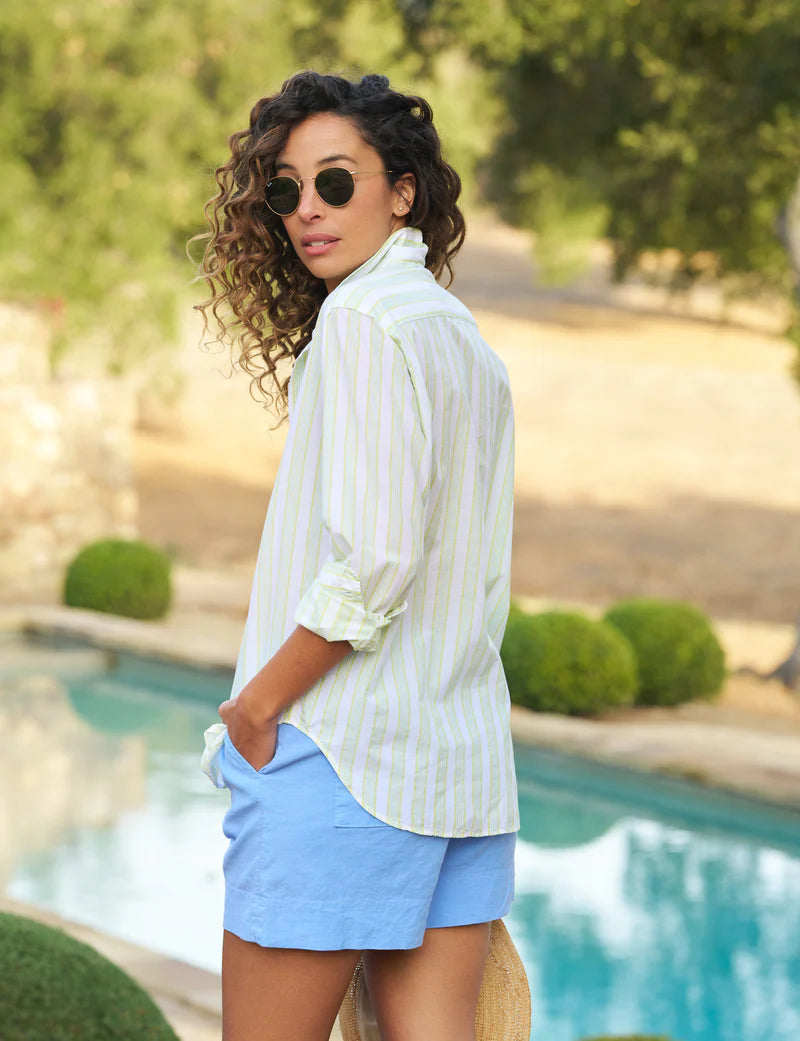 A person with curly hair in sunglasses and an Eileen S25 shirt made of Italian cotton by Frank & Eileen, paired with blue shorts, stands by a pool. Their hands are in pockets, exuding a relaxed fit vibe as they look back against a backdrop of trees and blurred greenery.