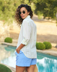 A person with curly hair in sunglasses and an Eileen S25 shirt made of Italian cotton by Frank & Eileen, paired with blue shorts, stands by a pool. Their hands are in pockets, exuding a relaxed fit vibe as they look back against a backdrop of trees and blurred greenery.