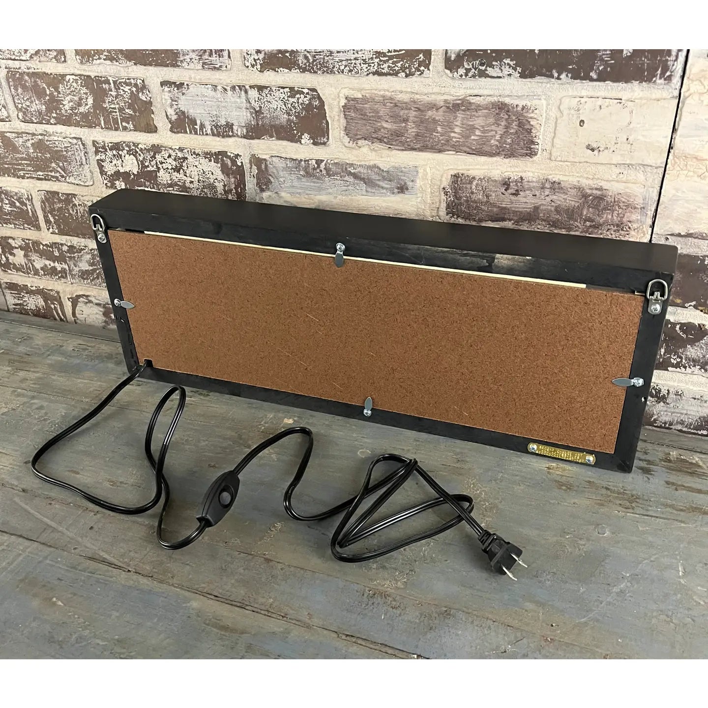 The Faire Lighted Sign features a rectangular design with corkboard back, black frame, and attached cord with switch and plug. Shown on a wooden surface against a brick wall.