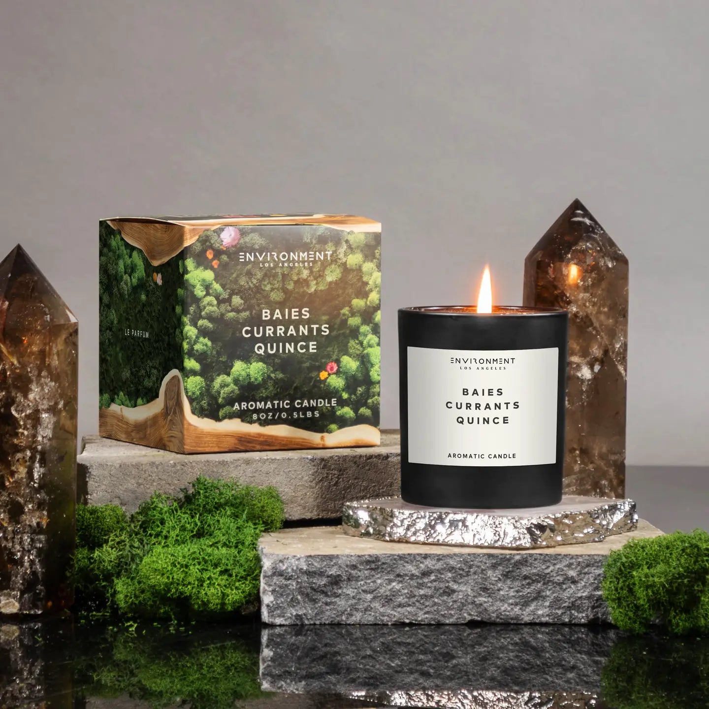 A lit Faire 8 OZ Candle With Lid labeled "Baies Currants Quince" rests on a stone platform next to its box, surrounded by green moss and two tall brown crystals, set against a gray backdrop—a true luxury scented candle experience.