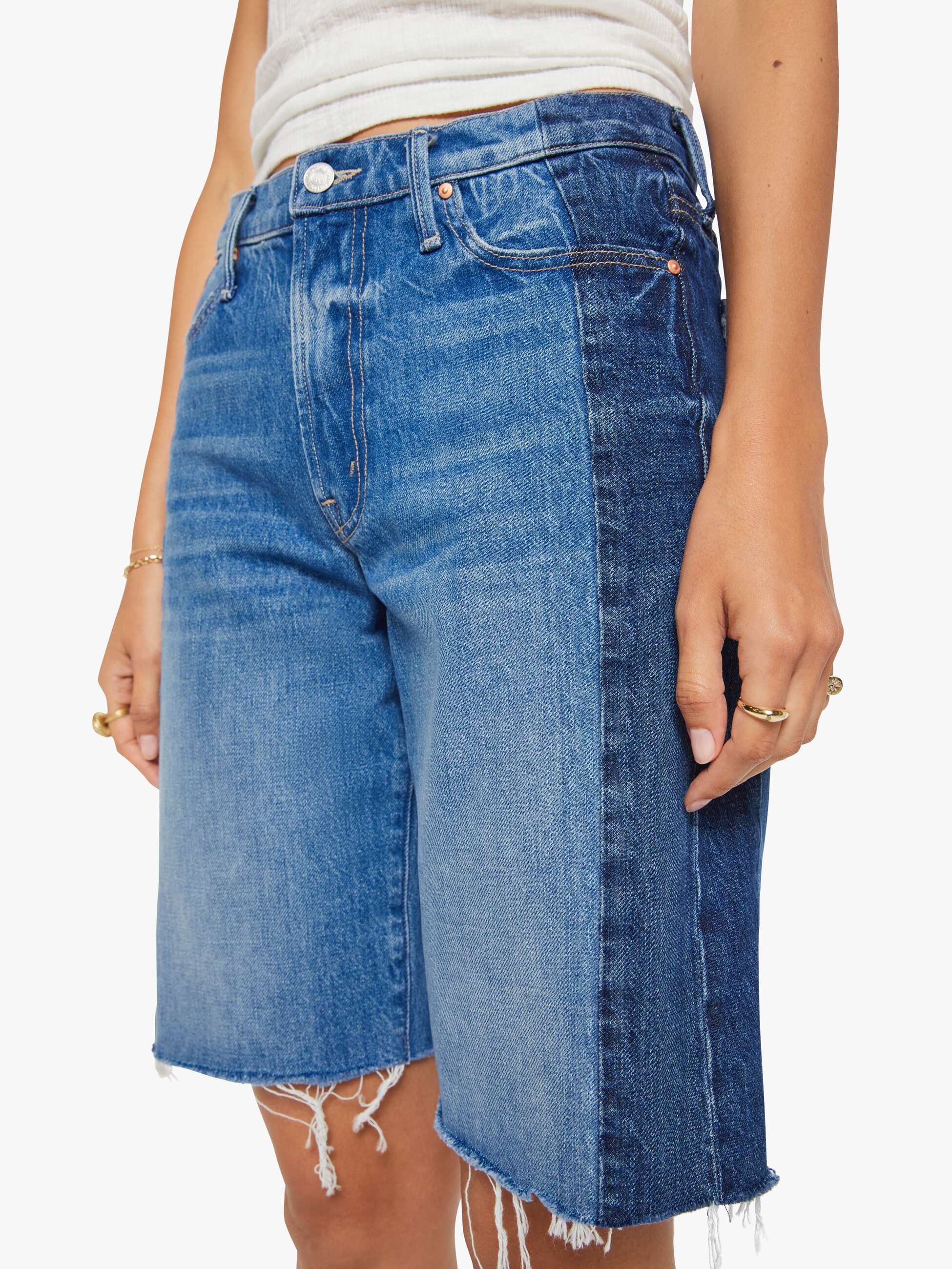 A person wears the Mother Mid Rise Maven Bermuda Fray shorts featuring a light blue front, dark blue sides, raw frayed hems, metal button and pocket details, paired with a white ribbed tank top for a stylish wide leg look.