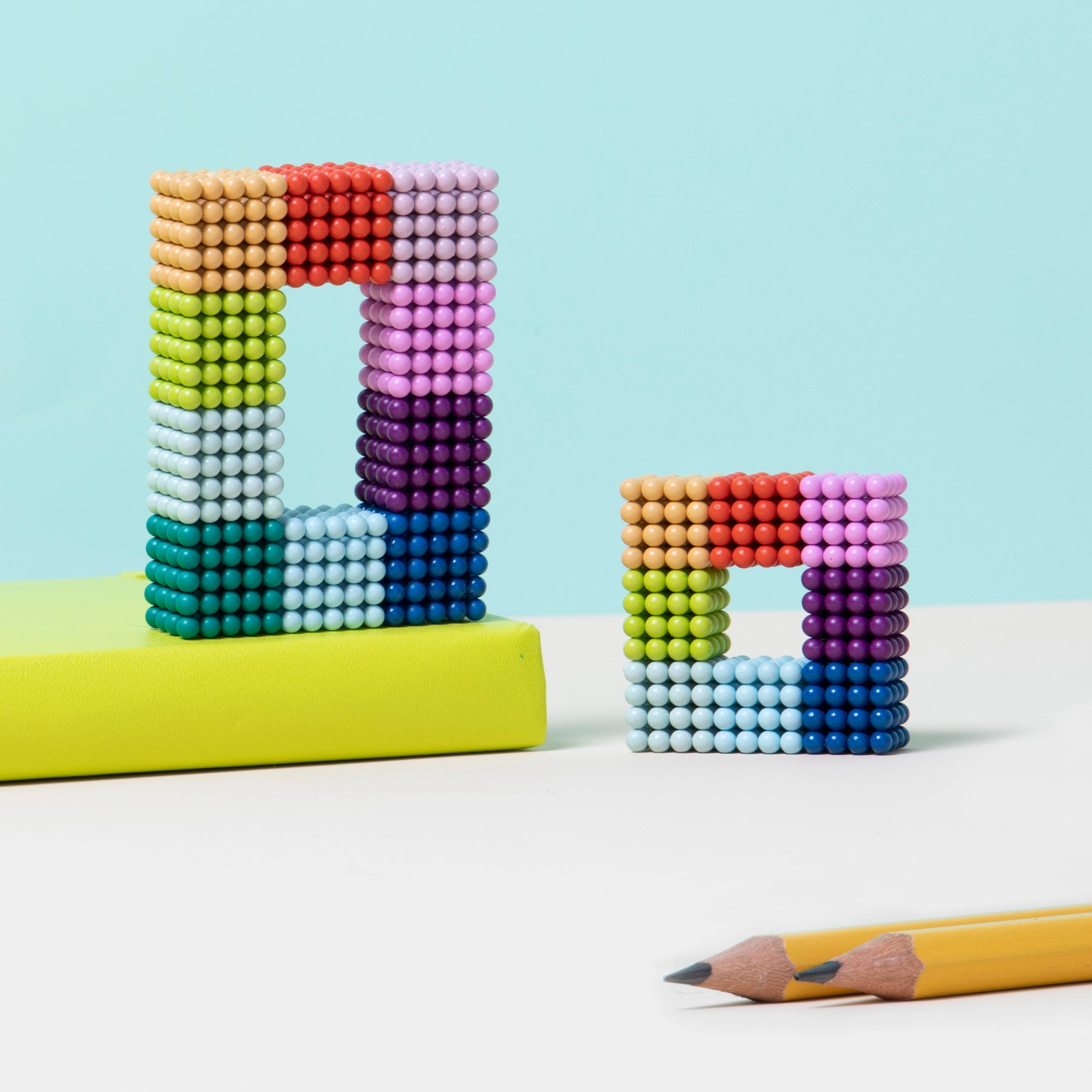 Two vibrant shapes made from Faire’s Speks- Matte Spectrum magnet balls sit on a white surface—one leans against a yellow book, with two sharpened yellow pencils nearby. This playful scene, set against a light blue background, offers stress relief.
