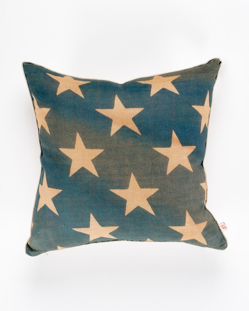 A Decorative pillow with a dark blue background featuring a pattern of large, beige stars. The &quot;Old Glory Stars 26x26&quot; pillow from Design Legacy has a square shape and a slightly frayed edge with visible stitching, perfect for adding a touch of Scottsdale Arizona charm to any bungalow.