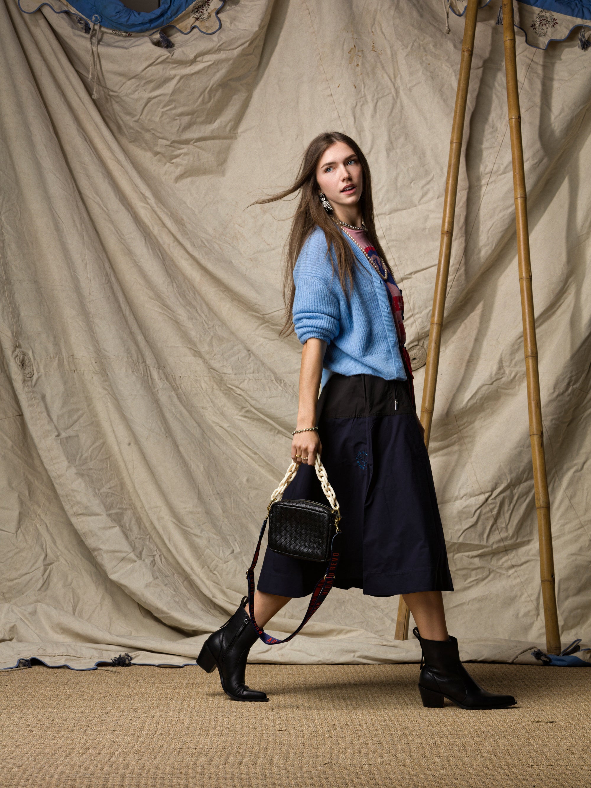 A woman with long brown hair walks in front of a beige draped backdrop, wearing a blue sweater, black ankle boots, and the Ping Pong Surf Club Le Vintage Sport Skirt. She carries a black chain-handle handbag.