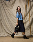 A woman with long brown hair walks in front of a beige draped backdrop, wearing a blue sweater, black ankle boots, and the Ping Pong Surf Club Le Vintage Sport Skirt. She carries a black chain-handle handbag.
