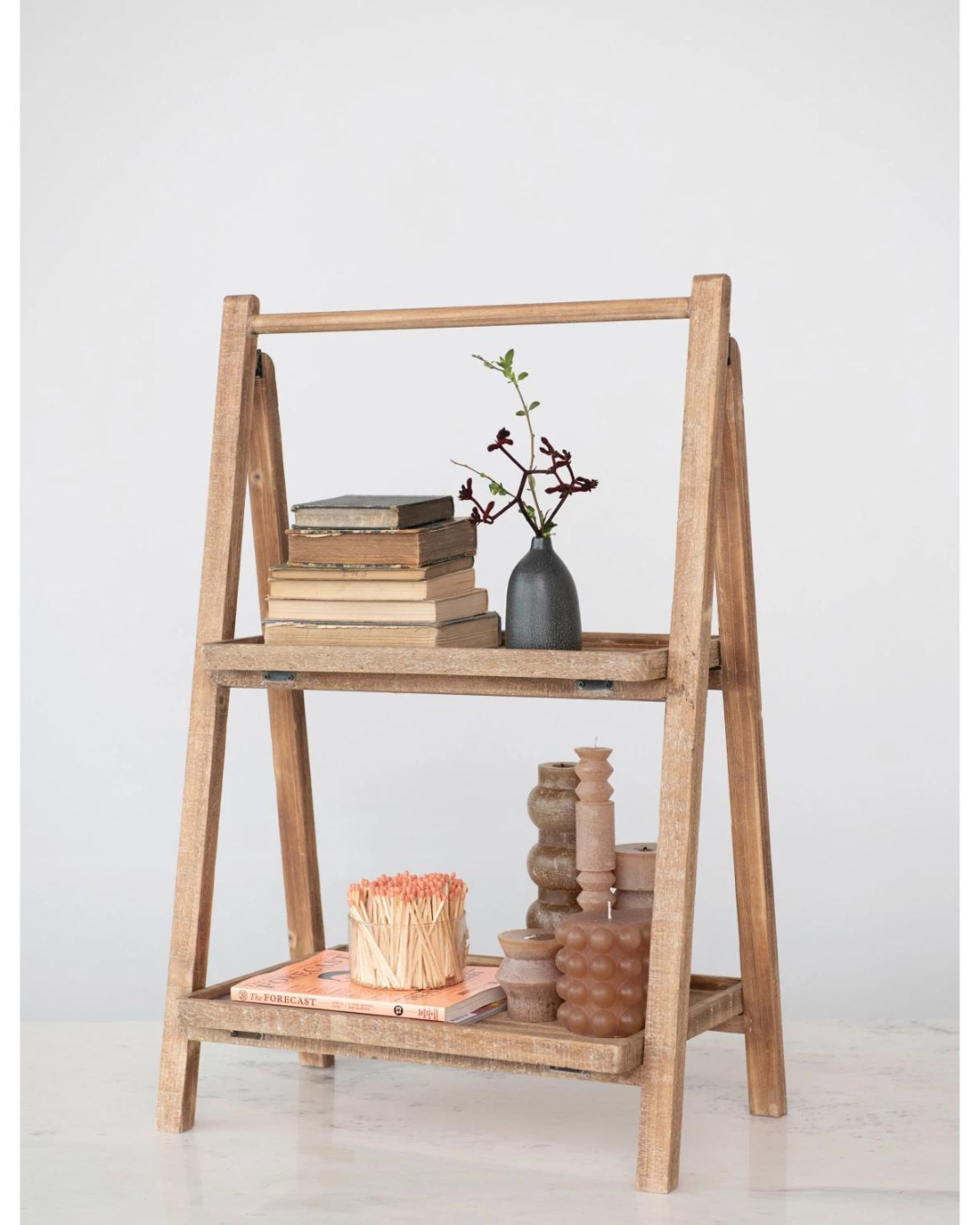 A Creative Co-op Two Tier Wood Folding Stand with a natural finish stands against a light-colored wall, holding folded books and a vase with branches on the top tier. The bottom shelf displays stacked books, a box of matches, and various candle holders and decorative items.