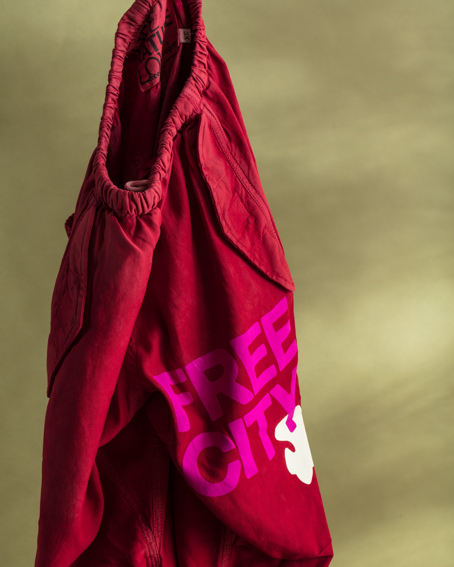 A pair of Flap/Snap LG MATTE/SATIN AirJump Pants from Free City (sparrow, LLC) is hanging against a greenish background. The pants feature the words &quot;FREE CITY&quot; in bold pink letters and a white dove graphic on the lower side. The waistband appears elastic, and the fabric looks casual and comfortable, boasting a custom sunfade wash for added style.