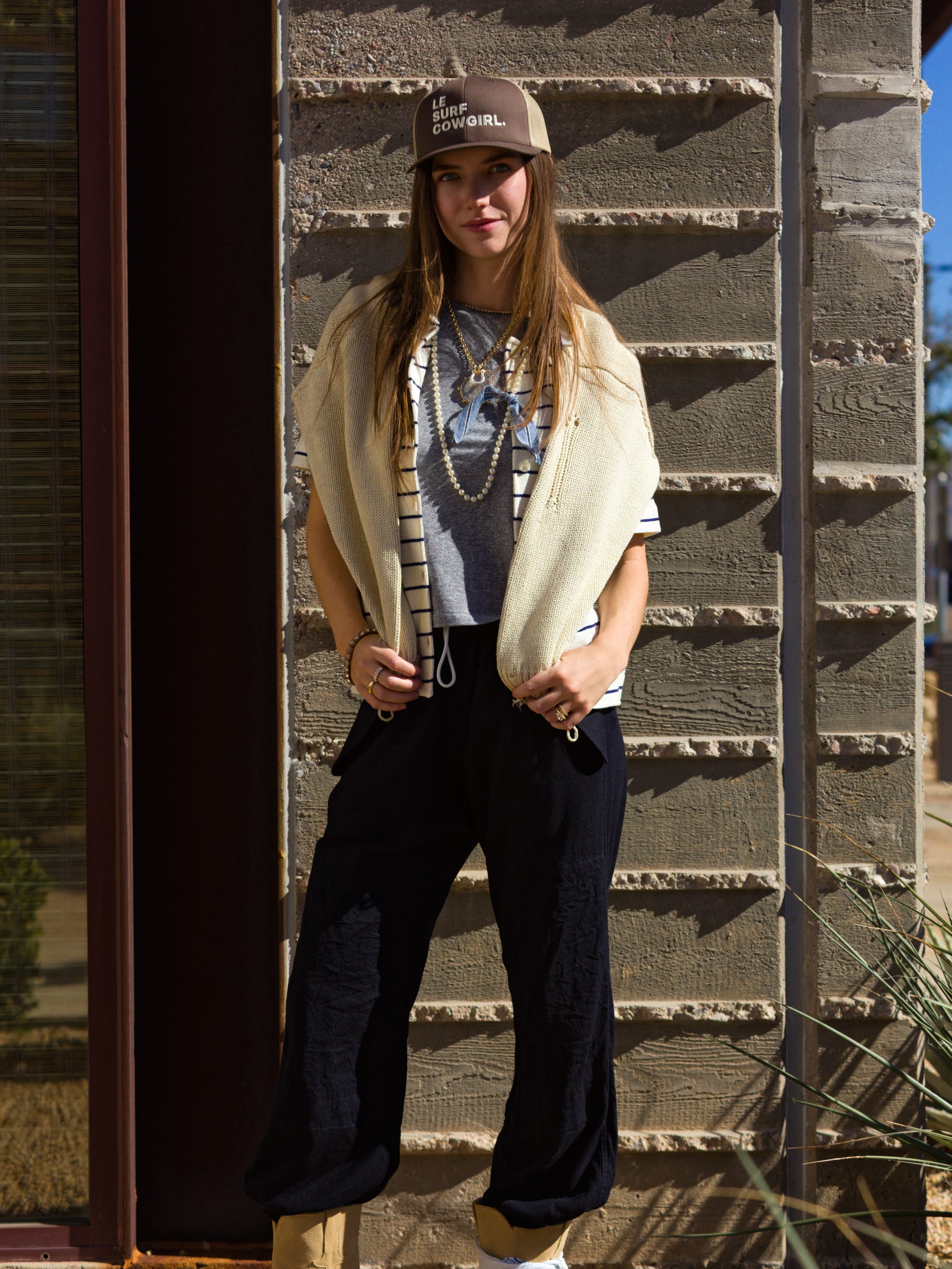 A woman stands outside against a textured wall, wearing the tan Le Surf Cowgirl Trucker by Ping Pong Surf Club, paired with a striped shirt, light sweater over her shoulders, dark pants, and tan boots. She faces the camera with a relaxed posture.