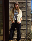 A woman stands outside against a textured wall, wearing the tan Le Surf Cowgirl Trucker by Ping Pong Surf Club, paired with a striped shirt, light sweater over her shoulders, dark pants, and tan boots. She faces the camera with a relaxed posture.