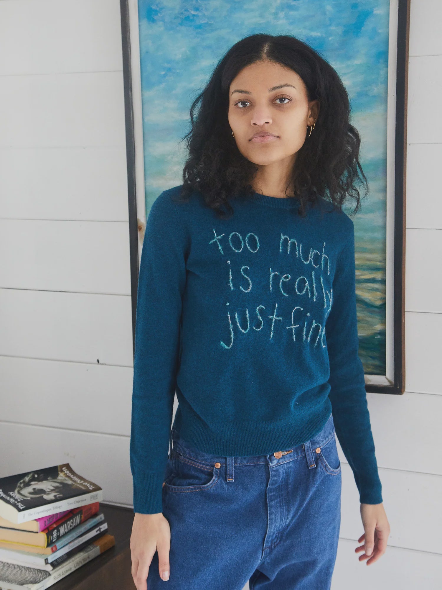 A woman with curly hair wears the Lingua Franca Cashmere Crew in teal, embroidered with “too much is really just fine,” standing indoors before a blue and white painting. A stack of books nearby evokes the laid-back Montauk spirit.