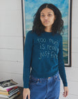 A woman with curly hair wears the Lingua Franca Cashmere Crew in teal, embroidered with “too much is really just fine,” standing indoors before a blue and white painting. A stack of books nearby evokes the laid-back Montauk spirit.