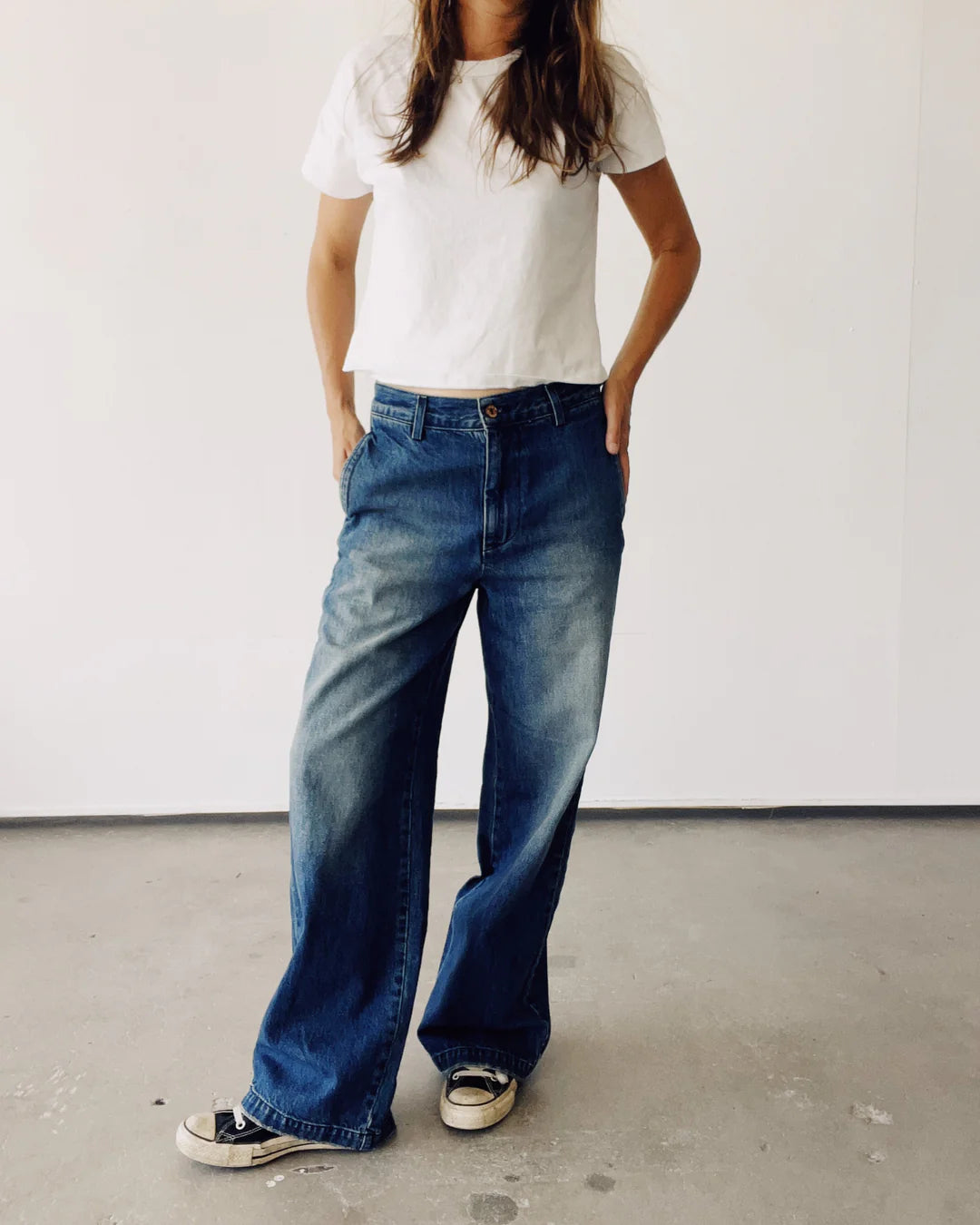 A person stands against a plain white background wearing a white T-shirt, The Buckle Back Trouser from Hey Gang with a slouchy fit, and black and white sneakers, with hands in pockets. Their face is not visible.