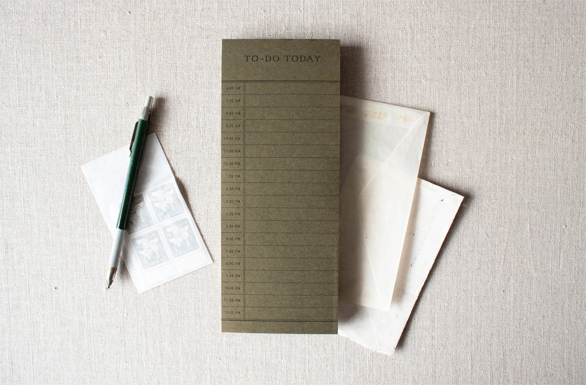 The Faire To Do Today Planner, a green daily notepad with hourly slots, is shown with a pen, postage stamps, and two envelopes on a light beige fabric surface.