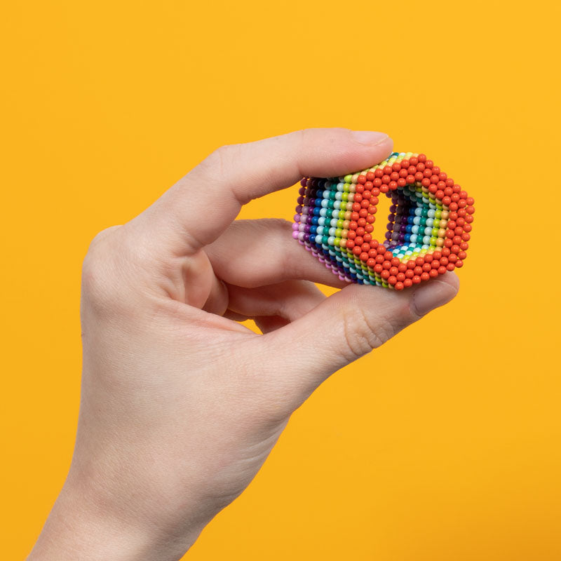 A hand holds the Faire Speks- Matte Spectrum, a hexagonal shape crafted from small, colorful magnetic balls, against a bright yellow background.