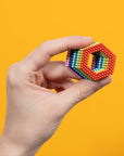 A hand holds the Faire Speks- Matte Spectrum, a hexagonal shape crafted from small, colorful magnetic balls, against a bright yellow background.