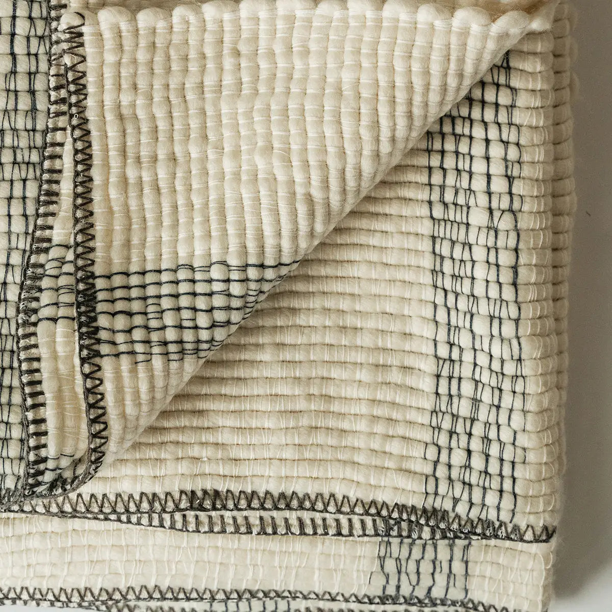 The Faire Cozy Stripe Throw is a folded, off-white wool-blend blanket featuring black stitched geometric lines. A turned-up corner reveals the stitching detail on both sides.