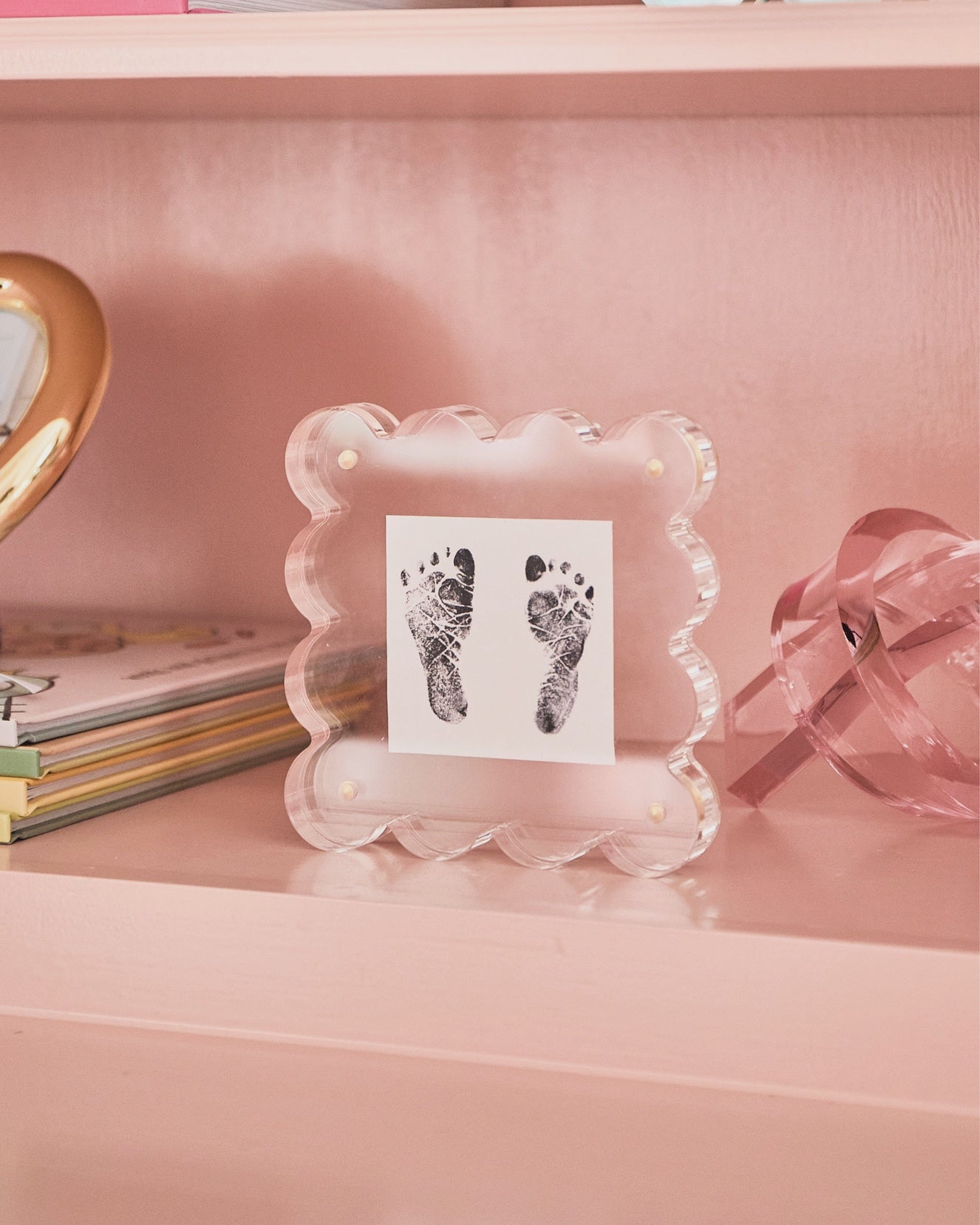 The Faire Acrylic Frame Mini, with a decorative scalloped design, showcases a black ink print of baby footprints on a pink shelf. Nearby, charming accents like a small book stack, heart-shaped object, and ribbon ornament complement the photo display.