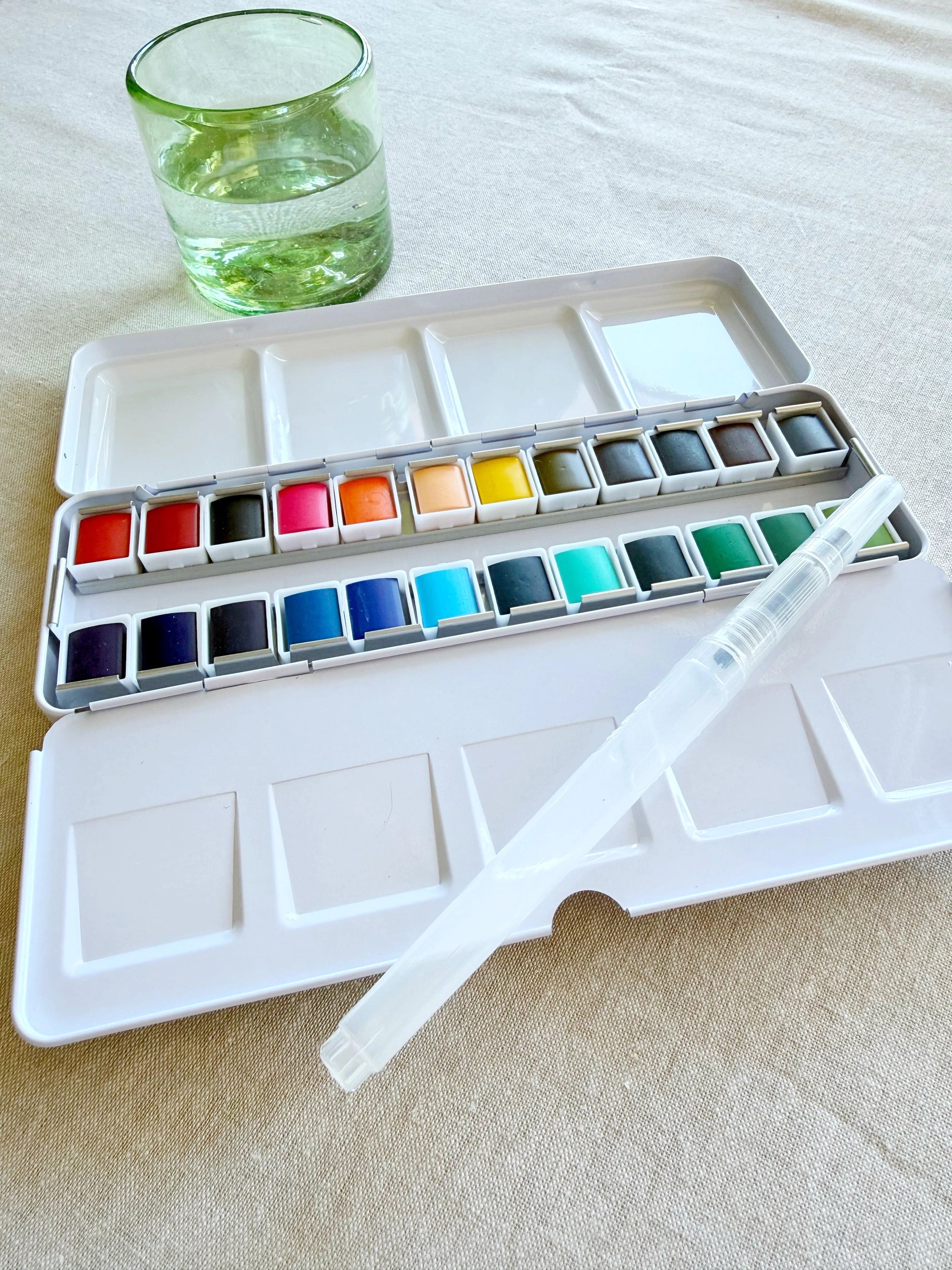 The Faire Watercolor Paint Set w/ Brush, plus an empty mixing palette and a glass of water, are placed on a beige tablecloth.