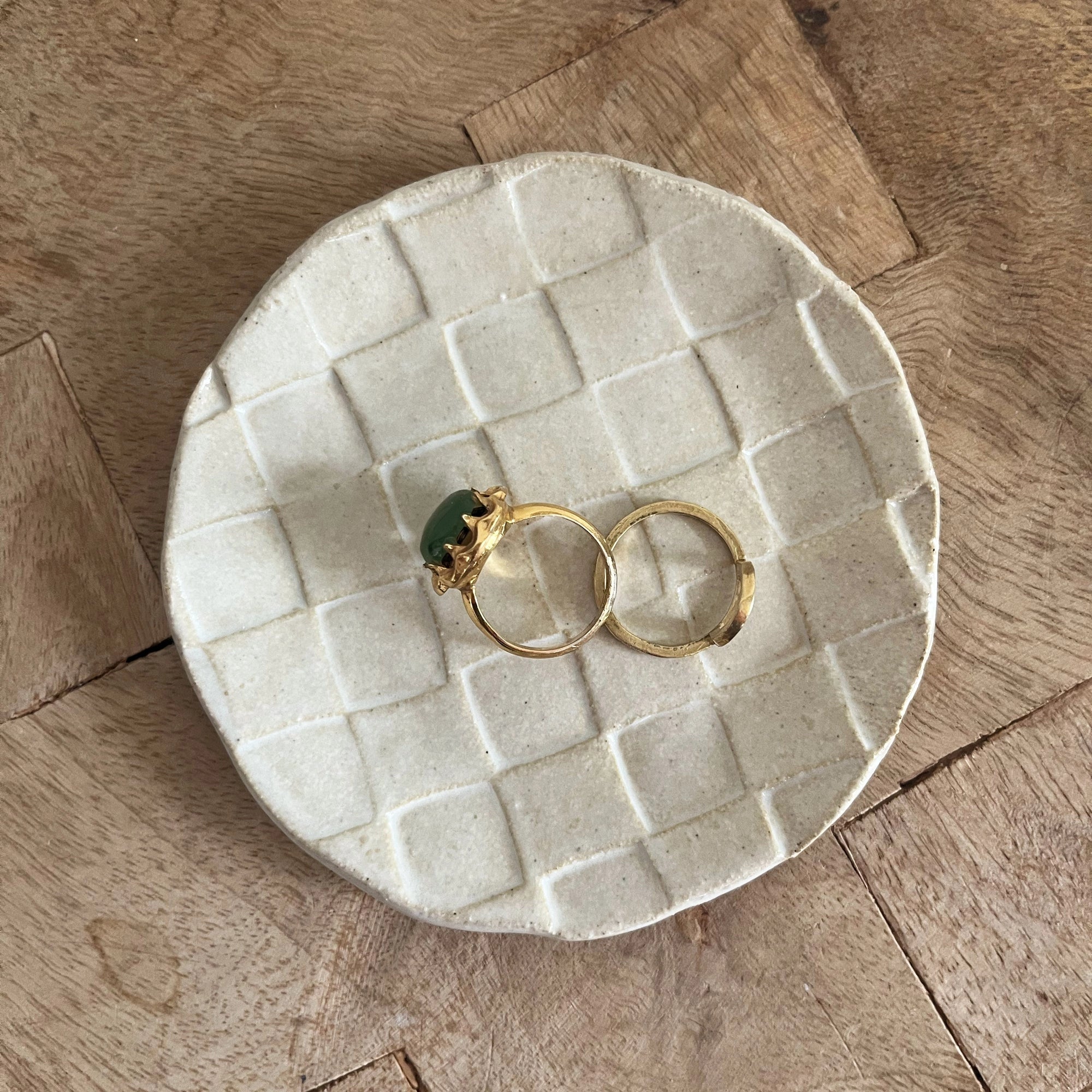 Two gold rings, one with a green stone, rest on Faire’s Ghost Checker Trinket Dish—a round, off-white glazed stoneware dish with a subtle checker pattern—displayed on a wooden surface.