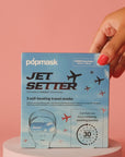 A hand holds a blue box of Faire's "Jet Setter" self-heating travel masks against a pink background; the packaging, adorned with airplane graphics, promises relaxation in 30 seconds.