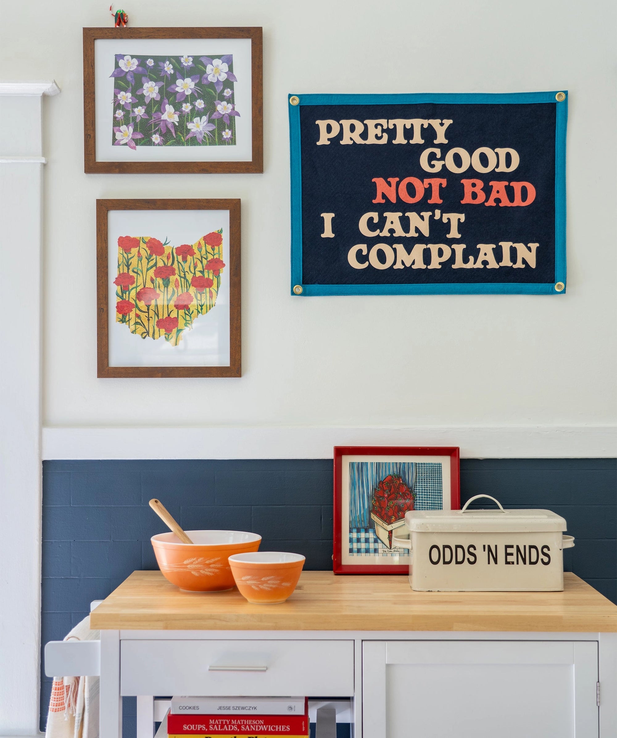 A small kitchen corner with orange mixing bowls, a cookbook, and a tin on a wooden cart. Above, framed wall art and an Oxford Pennant banner read “Pretty Good Not Bad I Can’t Complain.”.
