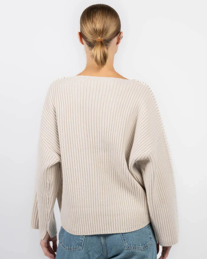A person with light brown hair in a low bun wears the Rag & Bone ingrid boat neck sweater in cream and blue jeans, facing away from the camera against a plain white background.