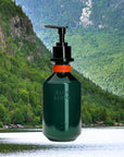 A dark green Faire Hand Wash Charlevoix Boreal dispenser sits before a lush forest, rocky cliffs, and a calm river—emphasizing its microbiome-friendly formula.