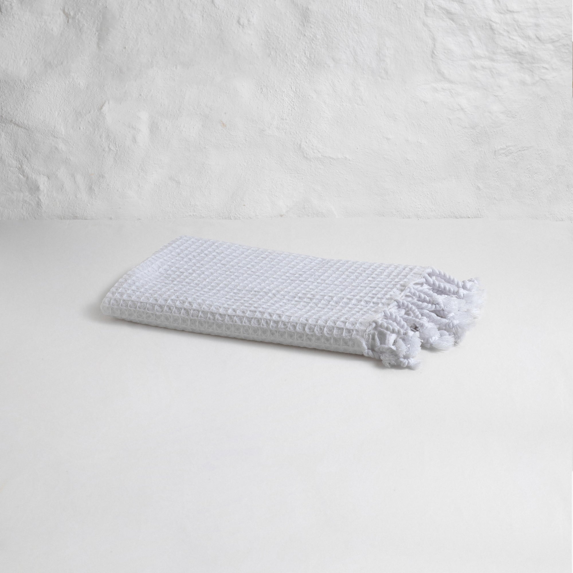 The Faire Waffle Hand Towel, crafted from white Turkish cotton with a waffle weave and tasseled edges, is displayed folded on a plain white surface against a textured white wall.