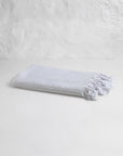 The Faire Waffle Hand Towel, crafted from white Turkish cotton with a waffle weave and tasseled edges, is displayed folded on a plain white surface against a textured white wall.