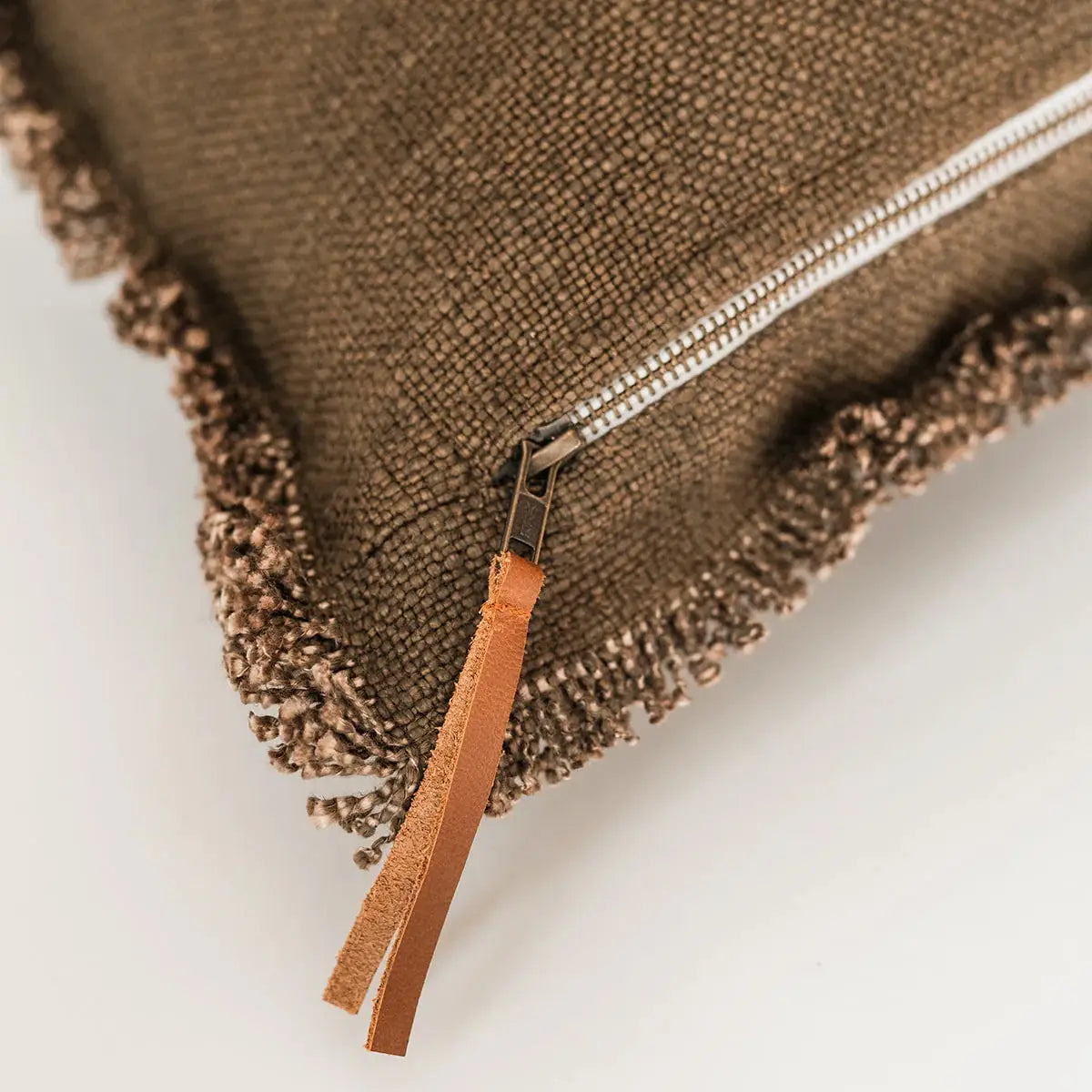 Close-up of the Luxe Linen Lumbar Pillow by Faire, a brown textured pillow with fringed edges, visible white zipper, and brown leather pull tab, shown on a plain white background.