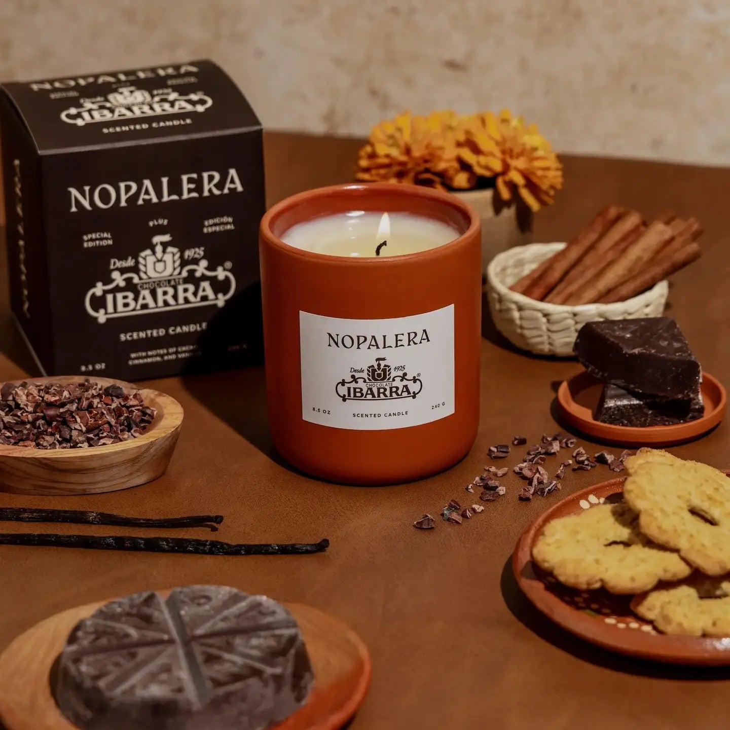 A lit Faire Nopalera Chocolate Candle sits on a table with cinnamon sticks, chocolate, vanilla beans, cookies, cacao nibs, and a marigold, evoking spiced hot chocolate. The candle’s box appears in the background.