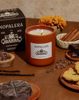 A lit Faire Nopalera Chocolate Candle sits on a table with cinnamon sticks, chocolate, vanilla beans, cookies, cacao nibs, and a marigold, evoking spiced hot chocolate. The candle’s box appears in the background.