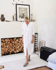 A person wearing the Rory Shirtdress S24 by Frank & Eileen and slippers stands in a living room featuring a white brick fireplace. Nearby, there's a stack of firewood, an amplifier, and some decorative pottery. A framed black-and-white artwork is displayed above the mantel.