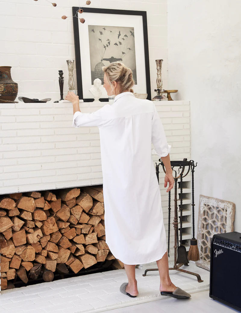 A person wearing a Rory Shirtdress S24 by Frank &amp; Eileen stands in front of a white brick fireplace filled with stacked logs. They are reaching for a framed picture above the mantle, which is adorned with various vases and candlesticks. An amplifier is visible on the floor.