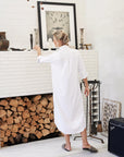 A person wearing a Rory Shirtdress S24 by Frank & Eileen stands in front of a white brick fireplace filled with stacked logs. They are reaching for a framed picture above the mantle, which is adorned with various vases and candlesticks. An amplifier is visible on the floor.