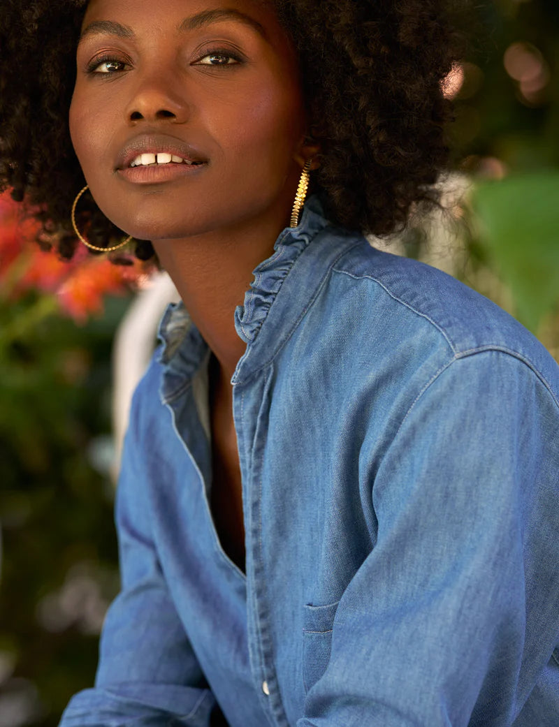 A woman with curly hair and large hoop earrings poses outdoors among green and red foliage, wearing the Frank &amp; Eileen Mary Classic Shirtdress- Ruffle.