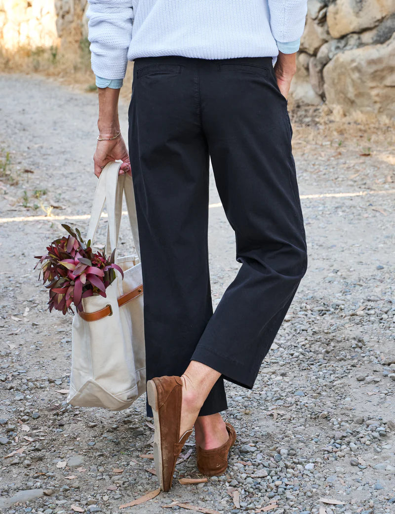 A person, seen from behind with only their lower body visible, walks on a gravel path wearing black Frank &amp; Eileen Westport Italian wide leg pants and brown loafers, carrying a canvas tote bag decorated with red foliage.