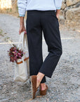 A person, seen from behind with only their lower body visible, walks on a gravel path wearing black Frank & Eileen Westport Italian wide leg pants and brown loafers, carrying a canvas tote bag decorated with red foliage.