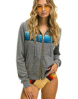 A woman with long wavy blonde hair wears yellow-tinted sunglasses, an Aviator Nation 5 Stripe Zip Hoodie in gray with blue stripes, and a tan bikini bottom with rainbow stripes, standing against a white background.