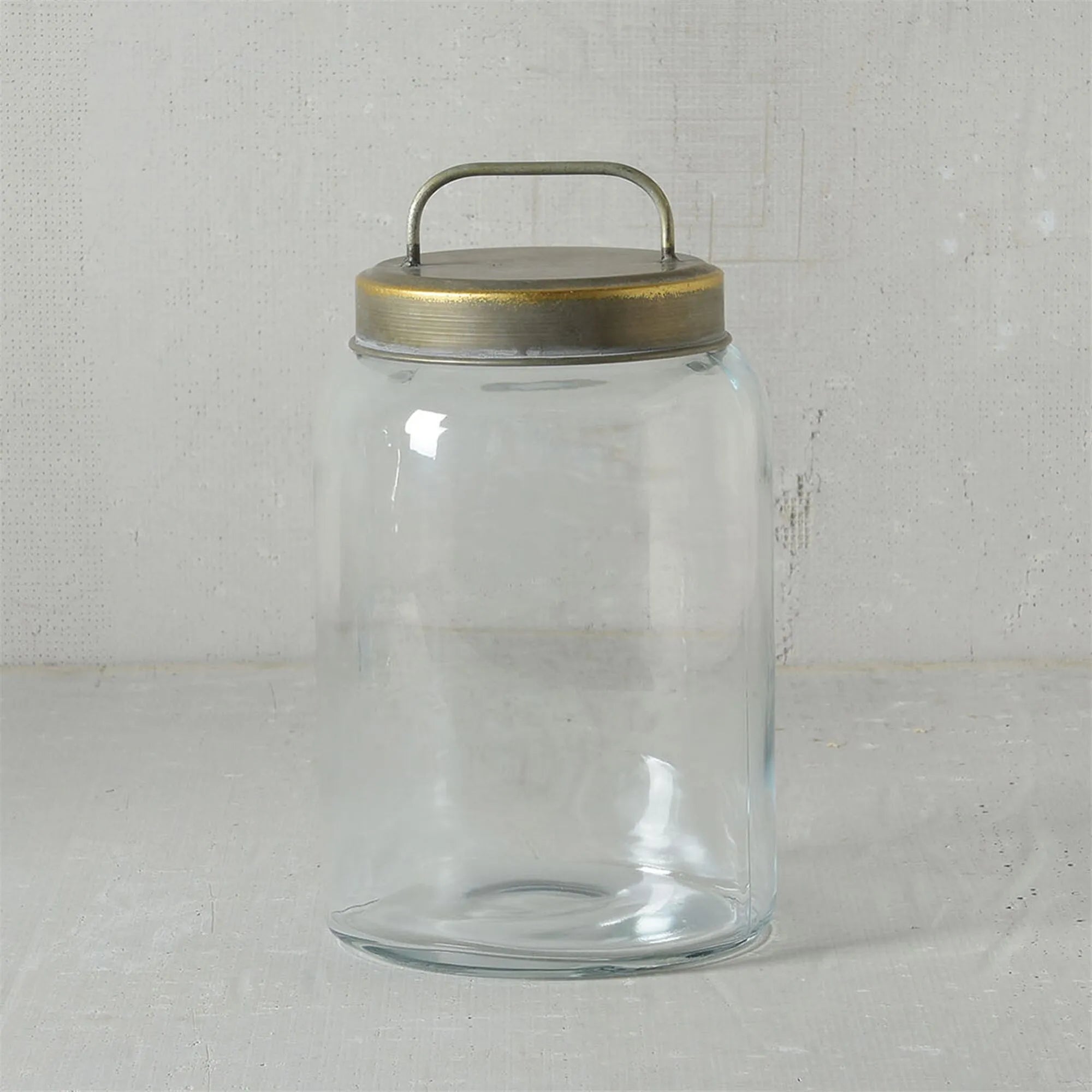 The Archer Canister Metal Lid by HomArt is a clear glass jar with a galvanized lid and small handle, shown on a light surface against a textured gray background. This empty cylindrical jar blends simplicity and functional style for perfect storage.