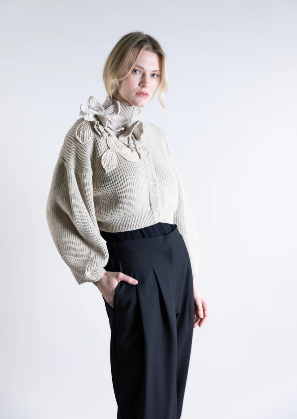 A woman wears the Psophia Funnel Neck Cardigan, a cream ribbed sweater with woven floral shoulder detail, paired with loose black high-waisted trousers, standing against a plain light background.