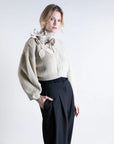 A woman wears the Psophia Funnel Neck Cardigan, a cream ribbed sweater with woven floral shoulder detail, paired with loose black high-waisted trousers, standing against a plain light background.