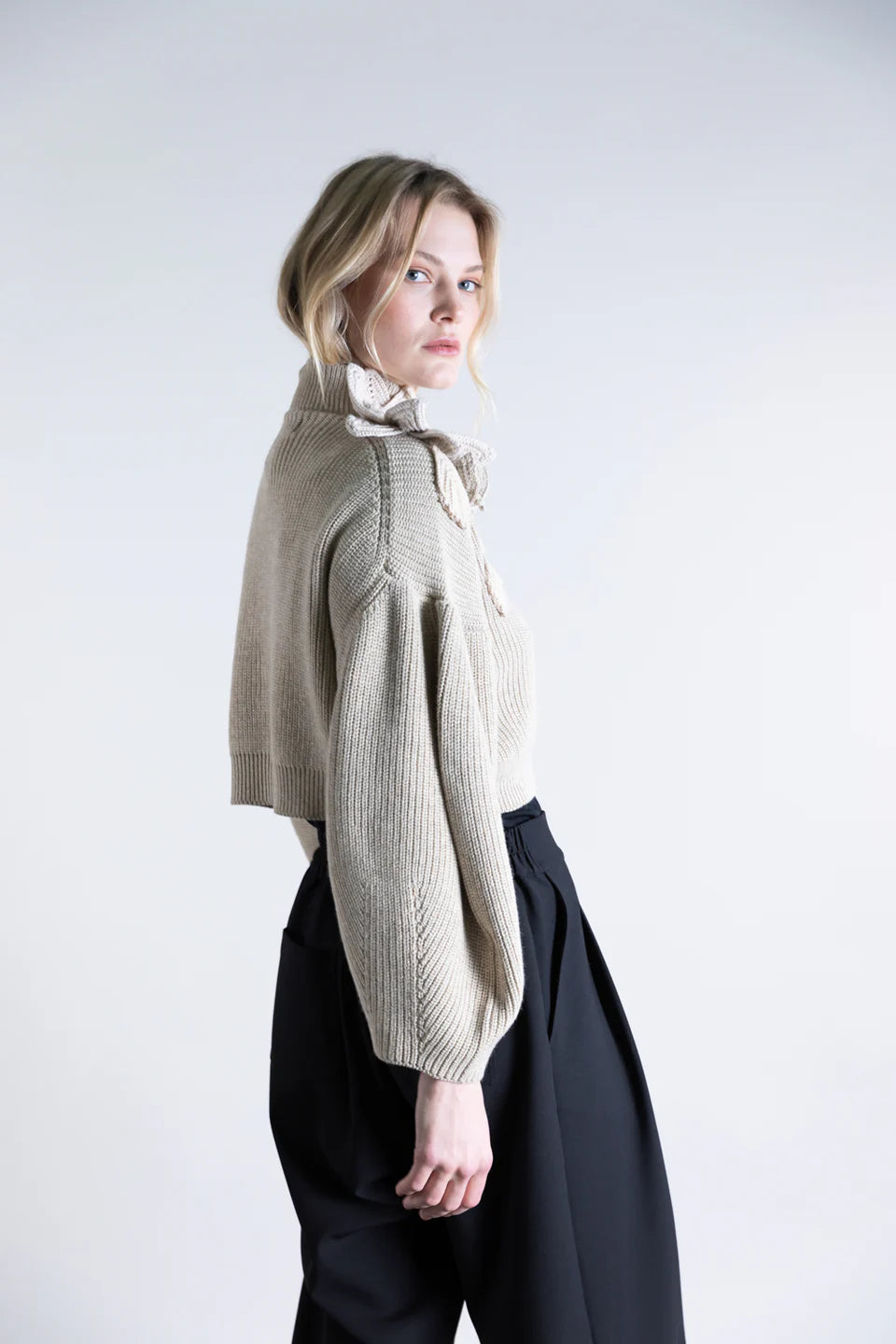 A person with blonde hair is wearing the Psophia Funnel Neck Cardigan, a beige, cropped style with bishop sleeves and a ruffled collar, paired with high-waisted black pants, standing sideways against a plain light background.