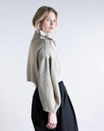 A person with blonde hair is wearing the Psophia Funnel Neck Cardigan, a beige, cropped style with bishop sleeves and a ruffled collar, paired with high-waisted black pants, standing sideways against a plain light background.