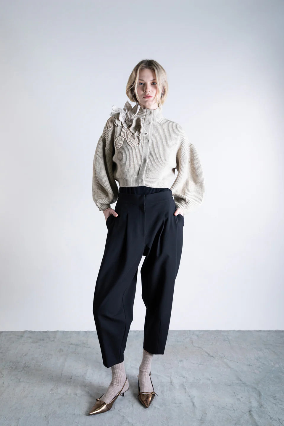 A woman stands against a plain background wearing the Psophia Funnel Neck Cardigan with woven floral shoulder detail, black wide-leg pants, beige socks, and metallic pointed-toe shoes, her hands in her pockets.
