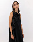 A woman with straight dark hair wears the Psophia 2784 Womens Dress, a sleeveless midi dress with a sculptural rosette detail on one side, standing against a plain white background.