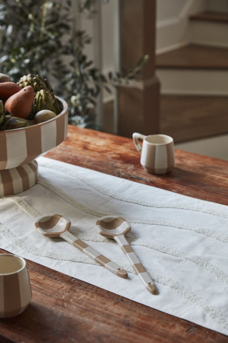 A wooden table with a white runner, two striped Botera Spoons, a bowl of fruit, and ceramic mugs. Blurred stairs and greenery appear in the background.